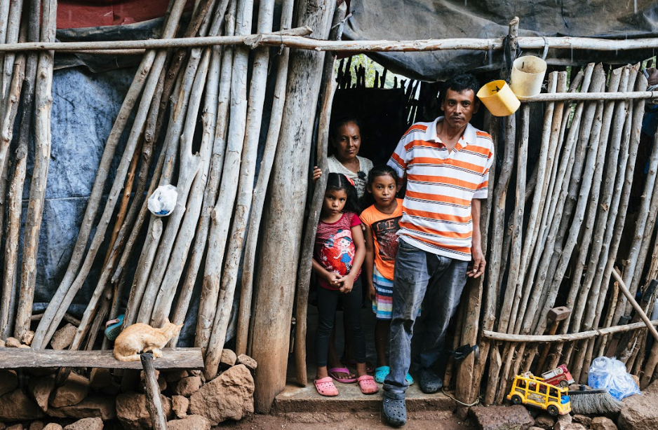 Hombre, su esposa y sus dos hijas parados en la puerta de su casa que está hecha de madera.