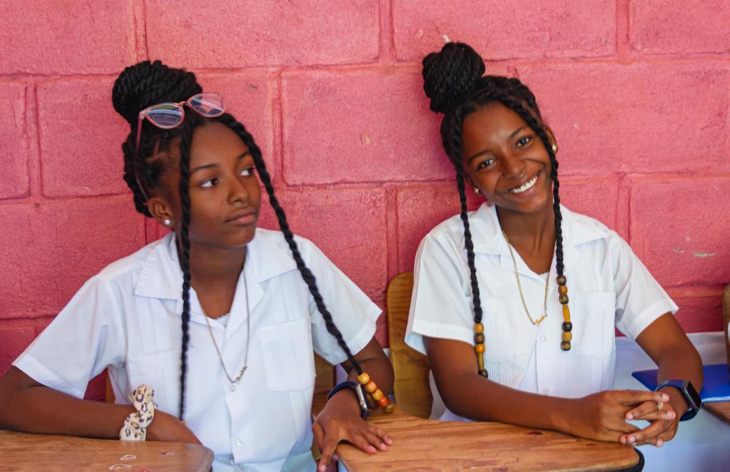 Dos jóvenes del municipio de El Limón, vestidas con uniformes blancos sentadas en pupitres de madera frente a una pared de ladrillo rosa. Ambas lucen trenzas largas; una de ellas sonríe, mientras la otra tiene una expresión pensativa, mirando hacia un lado.