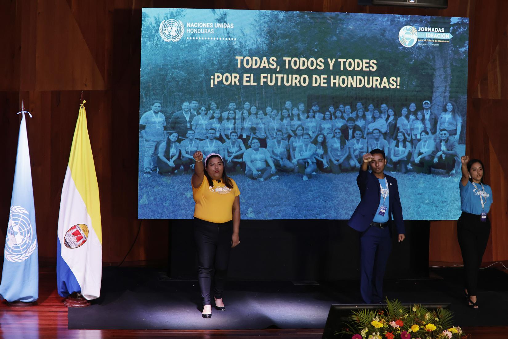 Nefer Mejía, Tyson García y Marlyn Brand, jóvenes participantes de las Jornadas de Ideación, presentan la visión conjunta en el escenario del Auditorio Alma Mater de la UNAH, vistiendo camisetas amarillas y azules que representan los colores de la Cumbre del Futuro.