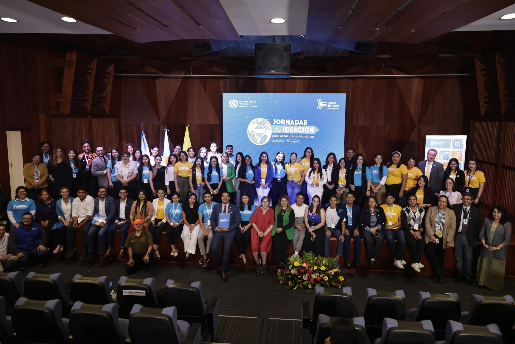 Jóvenes participantes de las Jornadas de Ideación para el Futuro de Honduras junto a mentores de las Agencias, Fondos y Programas de Naciones Unidas en el Auditorio Alma Mater de la Universidad Nacional Autónoma de Honduras, vistiendo camisetas azul cielo y amarillas, representando los colores de la Cumbre del Futuro.