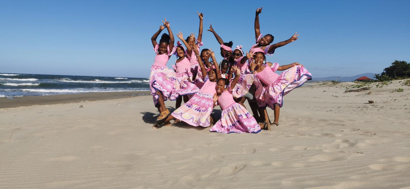 Grupo de niñas alzando sus brazos en la playa vestidas con trajes tradicionales coloridos, predominantemente en tonos rosas y lilas, con faldas amplias y detalles de patrones geométricos. De fondo, se puede ver el mar con olas suaves, una franja de arena y un cielo azul claro.