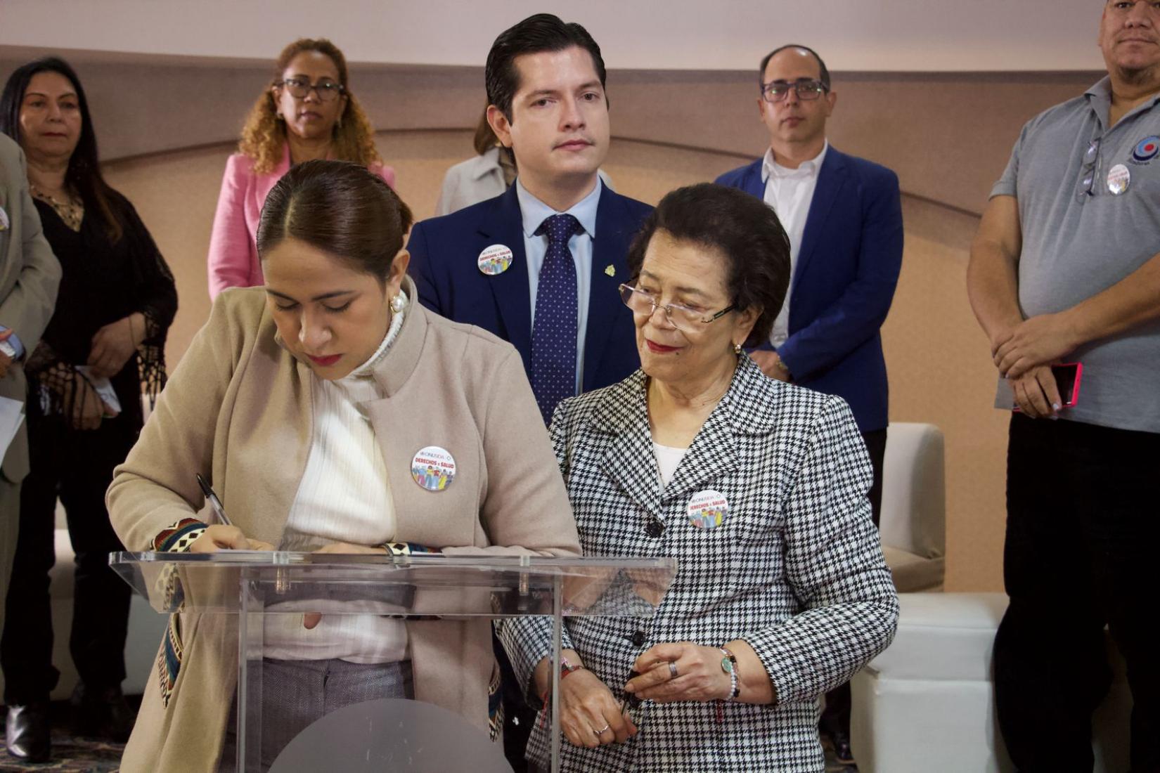 Angélica Álvarez, Ministra de la Secretaría de Derechos Humanos, viste un saco beige y camisa blanca, mientras que la Dra. Nerza Paz, Viceministra de la Secretaría de Salud, lleva un saco gris y usa lentes. Ambas firman el compromiso en la lucha contra el VIH/SIDA frente al público, desde un podio.