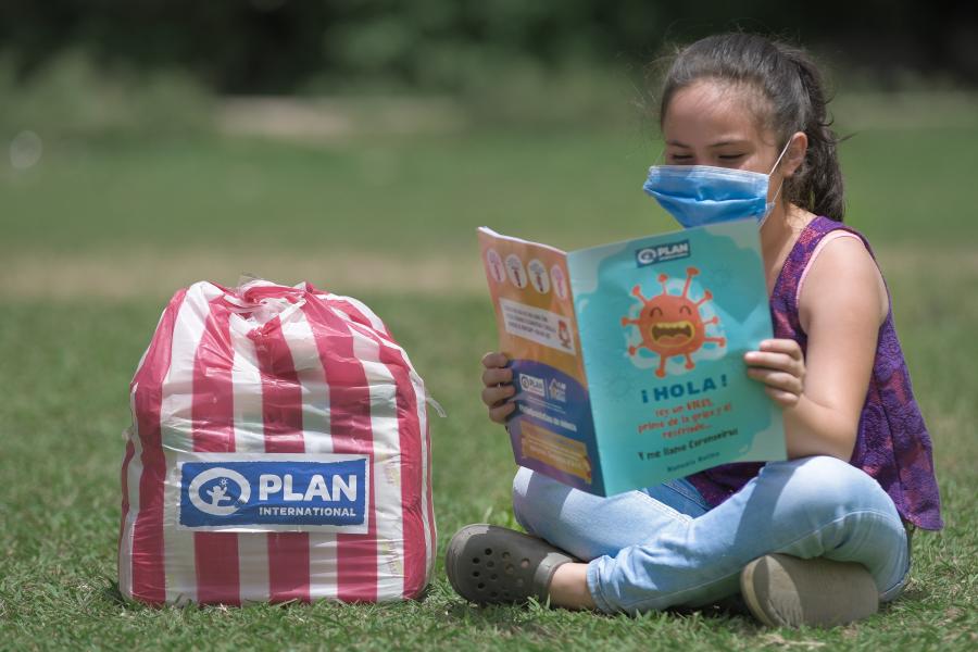 Niña sentada sobre grama junto a bolsa de alimentos de Plan Internacional Honduras leyendo folleto sobre prevencion COVID19