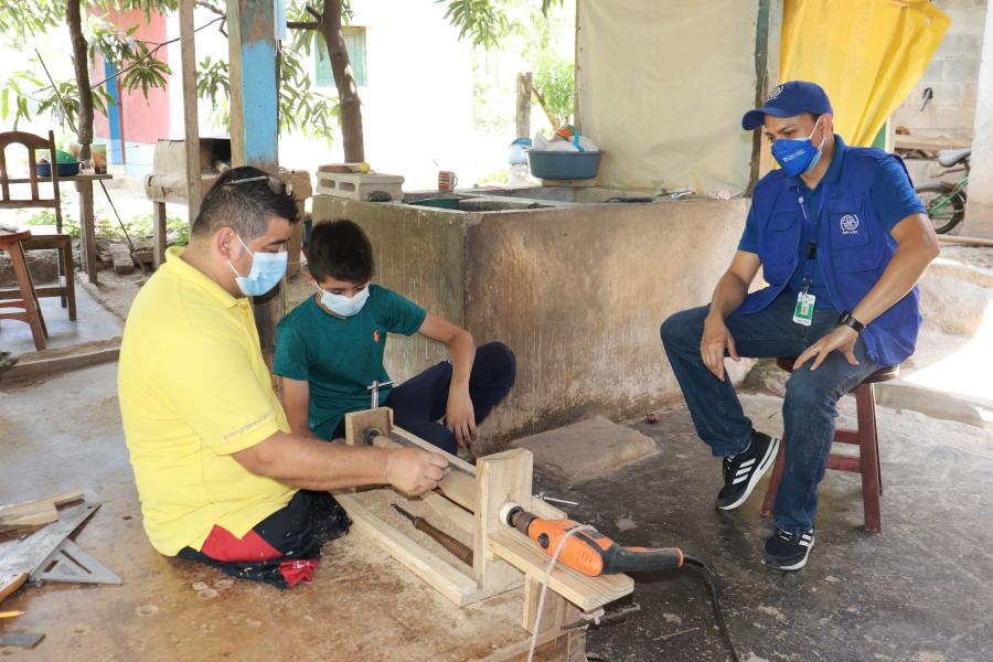 Un hombre y un menor trabajan con madera con el acompañamiento de un oficial de la OIM