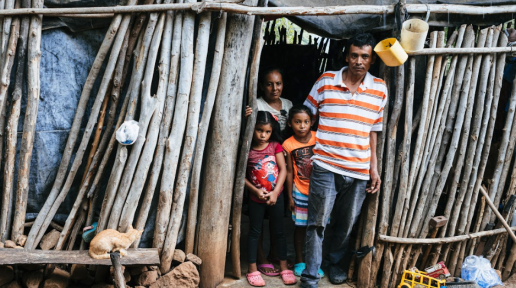 Hombre, su esposa y sus dos hijas parados en la puerta de su casa que está hecha de madera.