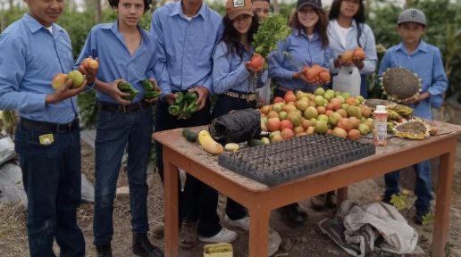 Grupo de jóvenes en un invernadero vestidos/vestidas con camisas de trabajo de color azul. Están sosteniendo diferentes productos agrícolas, como tomates, chiles y otras verduras recién cosechadas. Frente a ellos hay una mesa que también tiene una variedad de frutas y vegetales.