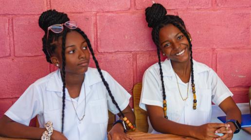 Dos jóvenes del municipio de El Limón, vestidas con uniformes blancos sentadas en pupitres de madera frente a una pared de ladrillo rosa. Ambas lucen trenzas largas; una de ellas sonríe, mientras la otra tiene una expresión pensativa, mirando hacia un lado.