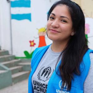 Danielle Alvarez ve a cámara con una leve sonrisa. Usa el pelo suelto con una camisa gris con las letras de ACNUR y un chaleco azul con las mismas.