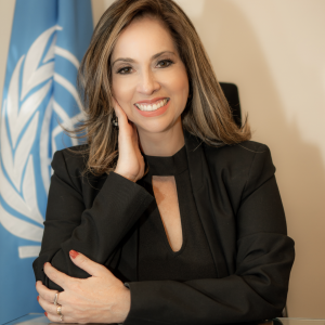 Claudia Valenzuela sentada frente a una bandera de Naciones Unidas sonriendo y viendo hacia enfrente