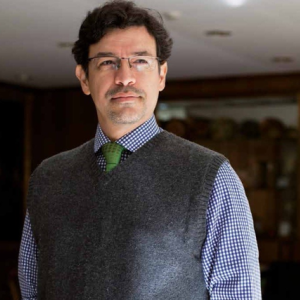 Fotografia de Elkin Velasquez, hombre de mediana edad, utilizando un chaleco de sueter gris sobre una camisa de cuadros y corbata verde con una expresión neutral viendo hacia delante