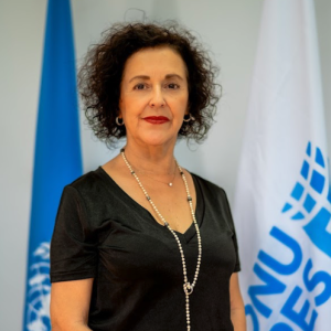 Margarita ve a cámara con una expresión suave, parada frente a las banderas de ONU y ONU Mujeres