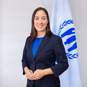 Kathryn Lo, frente a bandera de ACNUR usa un traje azul y sonríe a cámara